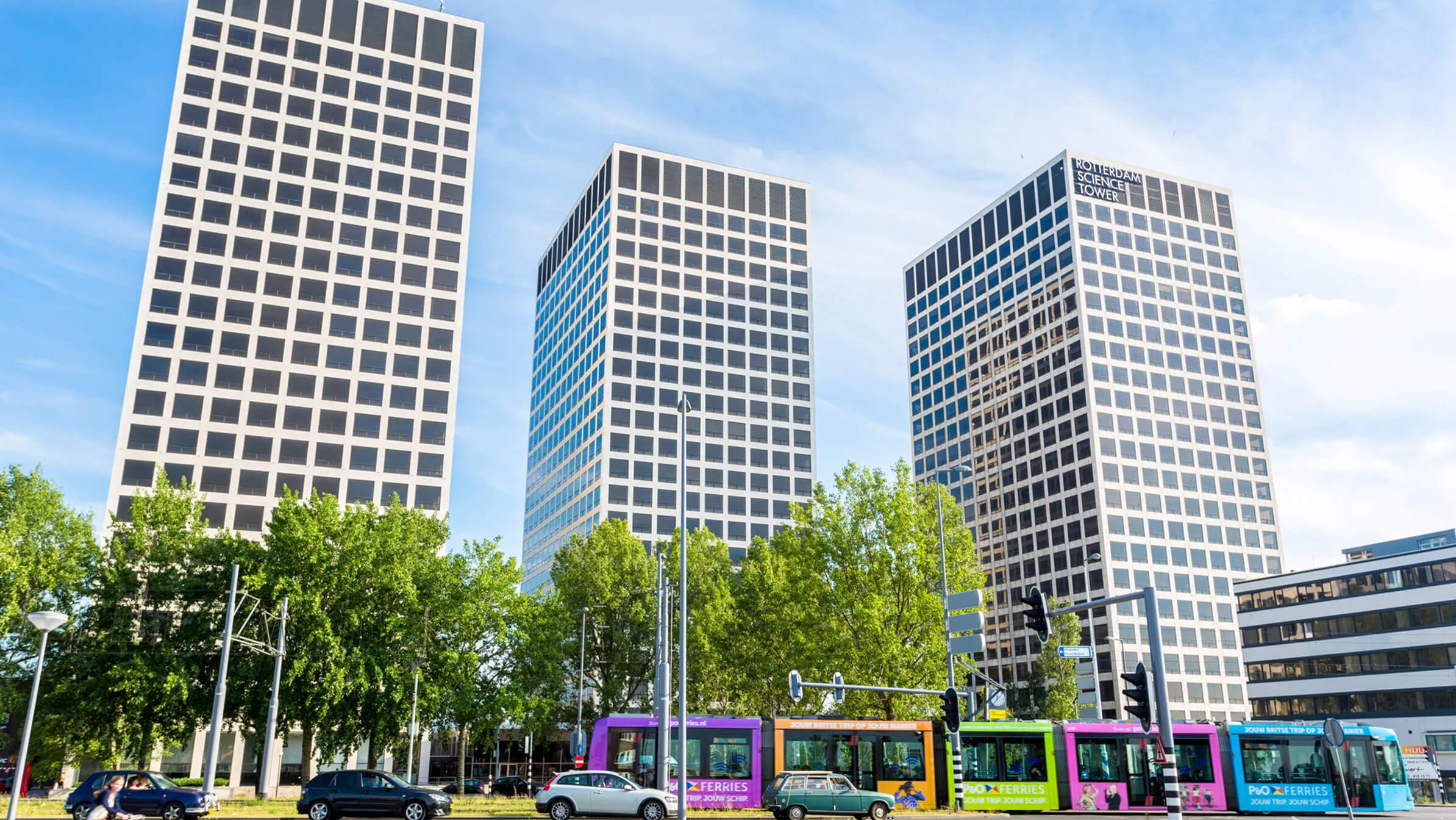 Drie hoge kantoorgebouwen met een tram en auto's ervoor, bomen op de voorgrond onder een blauwe lucht.