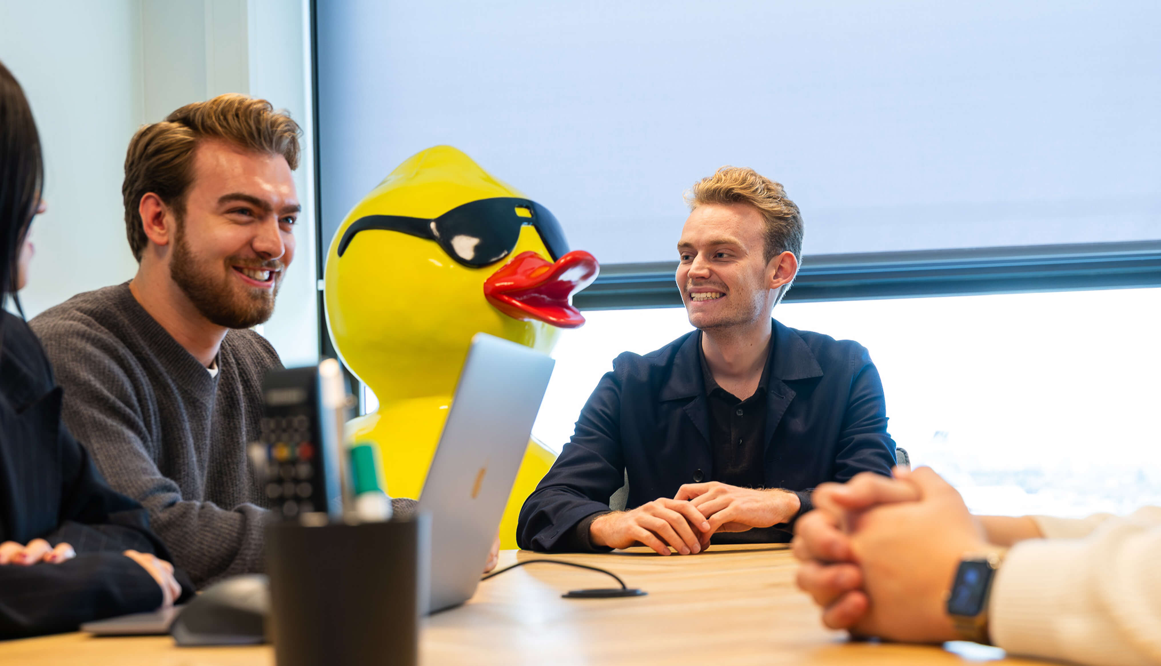 Twee lachende mannen zittend aan een tafel met een laptop en een groot, geel badeendbeeld met zonnebril in een kantooromgeving.
