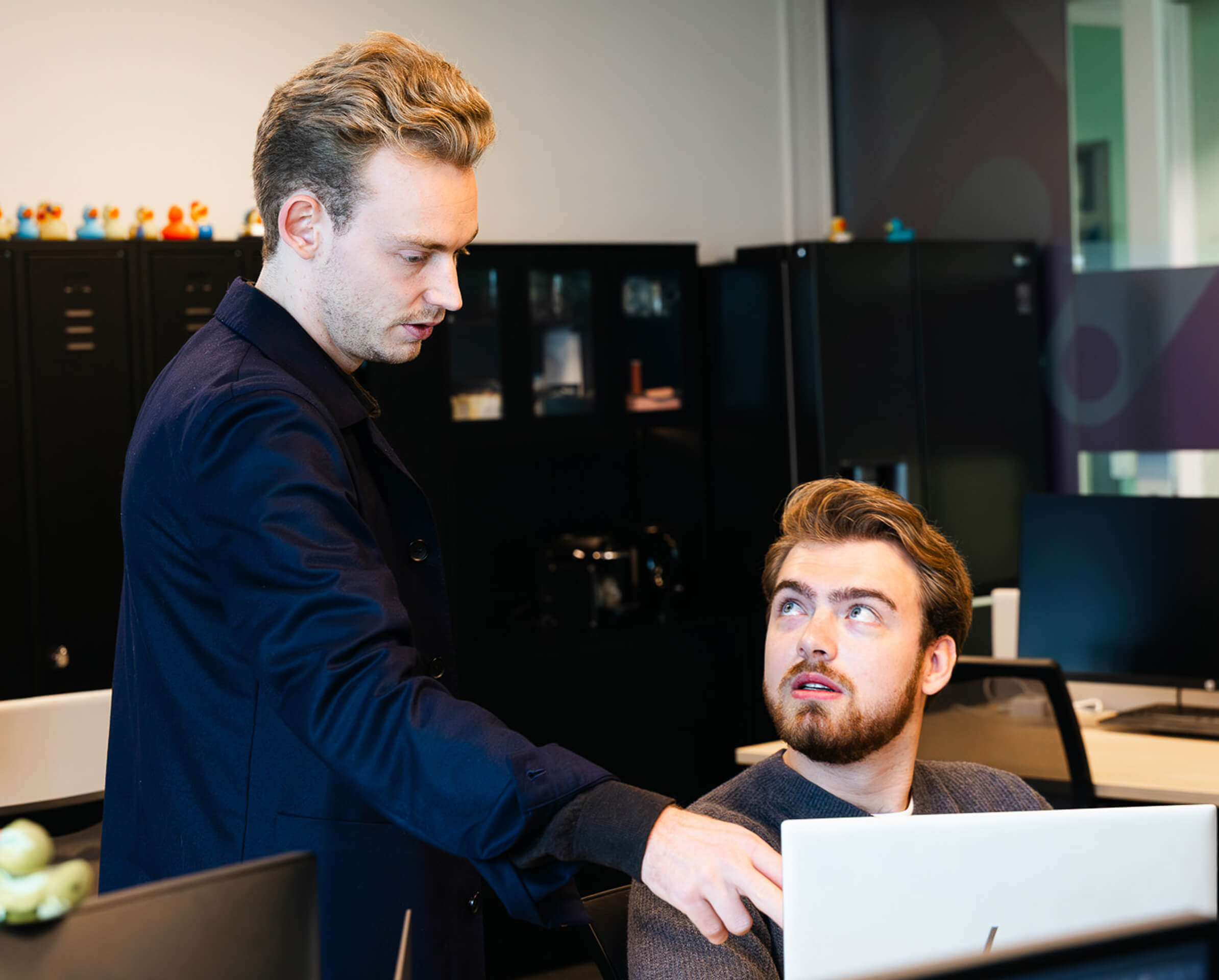 Twee mannen in een kantoorruimte, waarbij de staande man naar iets op het computerscherm van de zittende man wijst.
