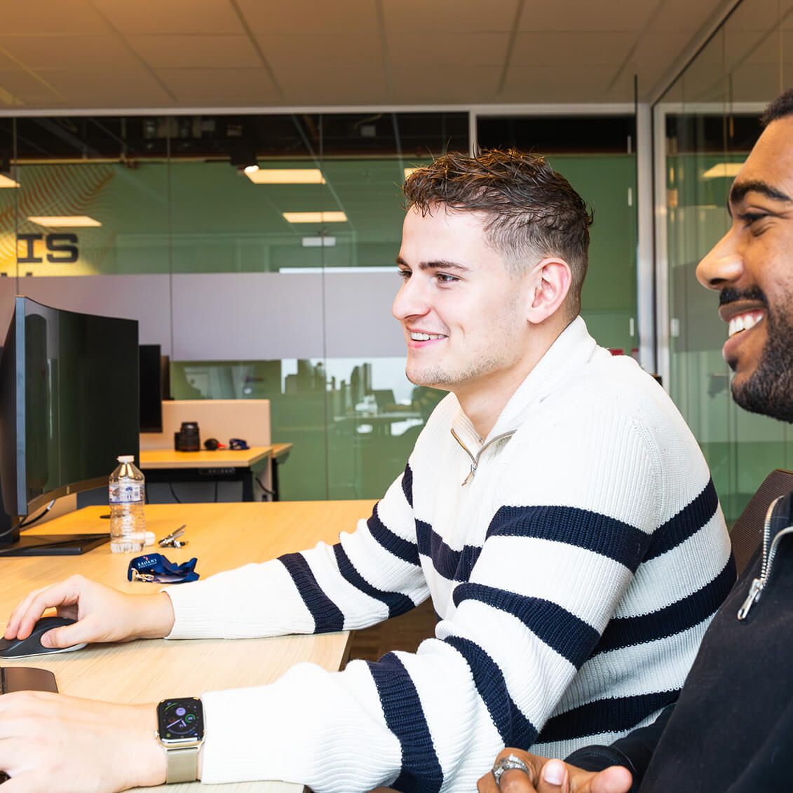 Twee mannen lachen terwijl ze samenwerken aan een bureau met een computer in een kantooromgeving.
