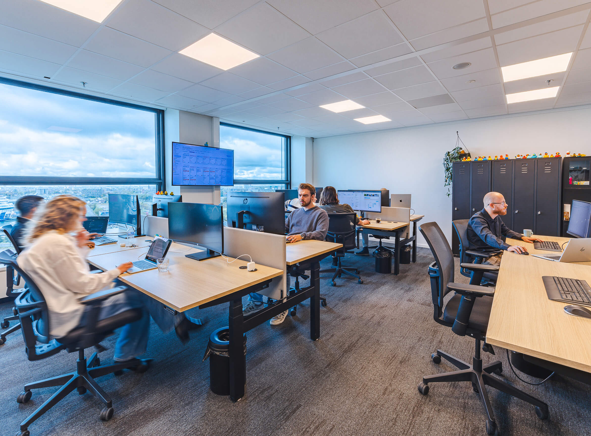Medewerkers werken op computers in een moderne kantoorruimte met grote ramen en een wandmonitor.