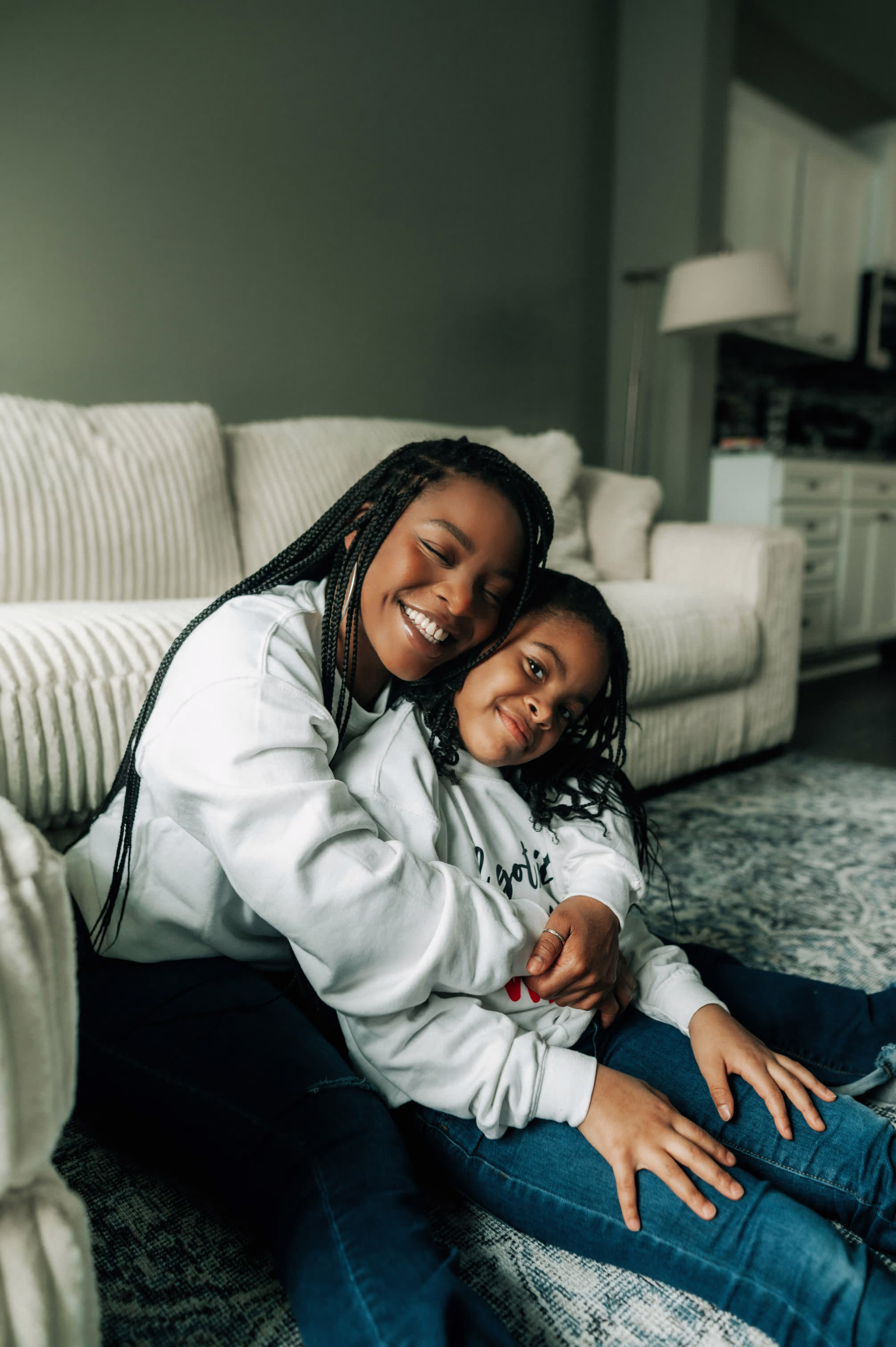 Smiling woman hugging a young girl while sitting on the floor in a living room with a beige sofa and lamp in the background.