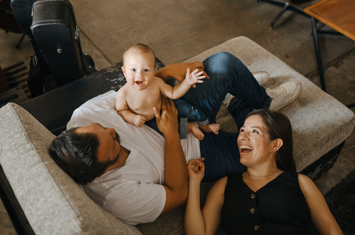 Smiling baby lying on a man's chest on a couch while a woman lies beside them looking happily at the baby.