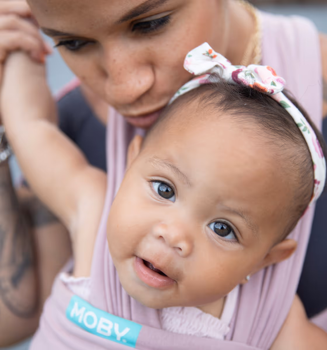Close-up of a baby with a floral headband looking at the camera, held closely by an adult wearing a pink sling.