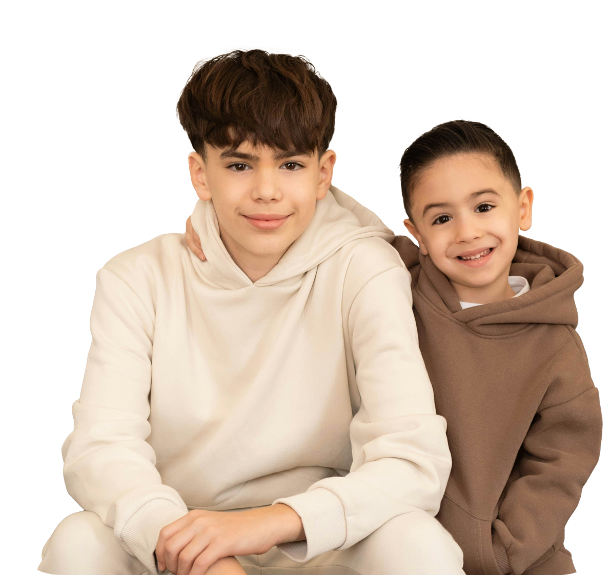 Two smiling boys sitting close together, one with brown hair wearing a cream hoodie, the other younger in a brown hoodie.