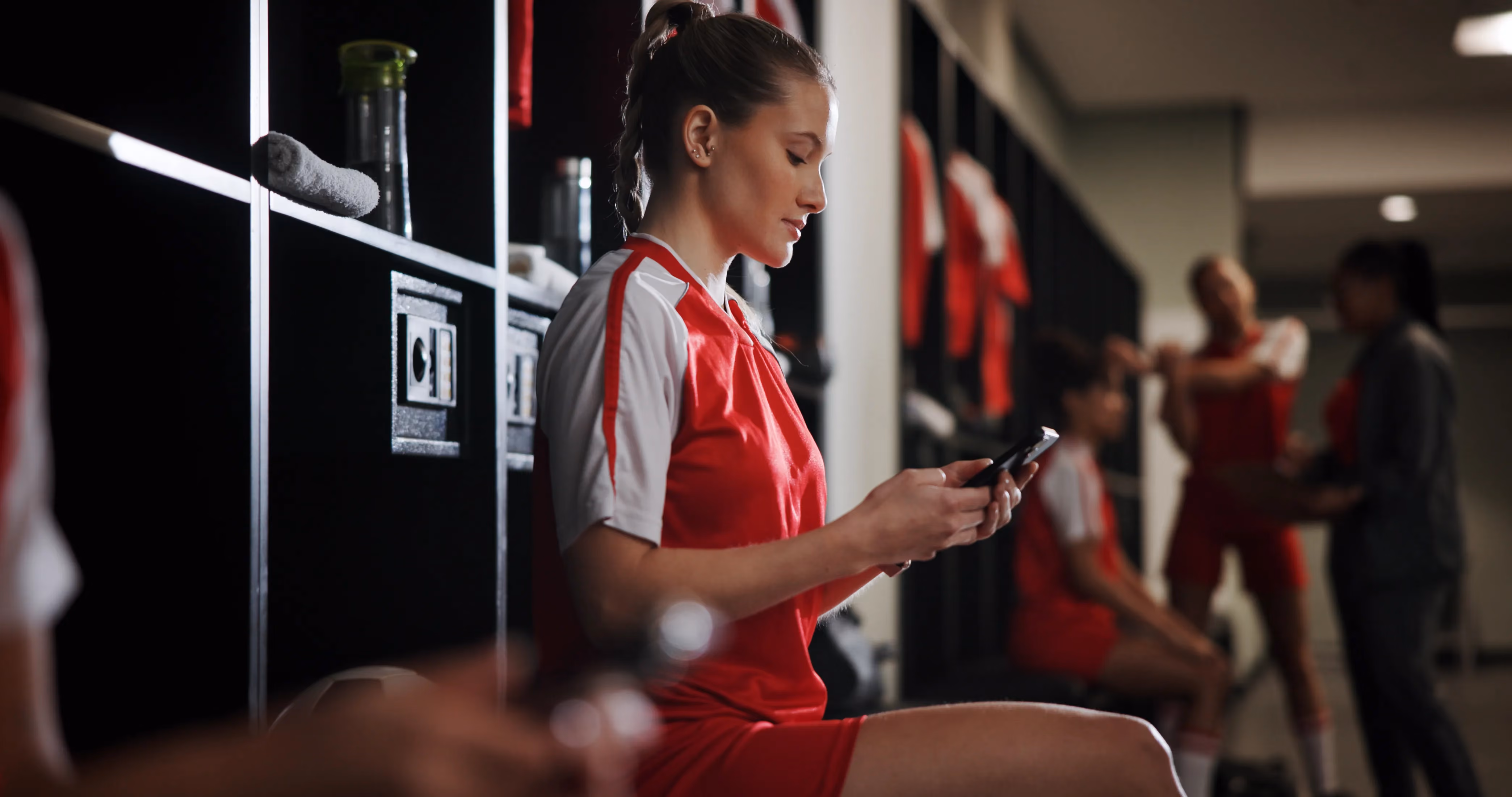 A female athlete in a red and white uniform sits in a locker room, focused on her phone while teammates talk in the background.