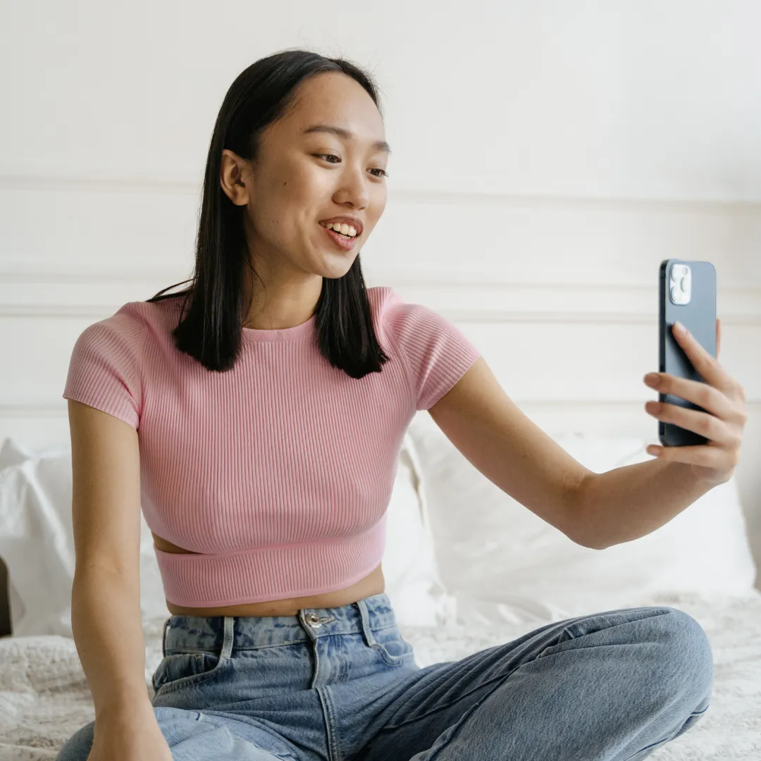 A young woman in a pink top and jeans sits on her bed, smiling as she records a video on her smartphone.