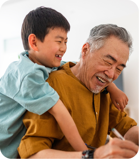 Joyful photo of a grandfather in a mustard shirt smiling and writing while his grandson in a light blue shirt hugs him from behind.