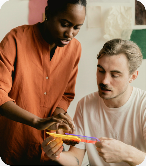Two people collaborating: a woman in an orange shirt leans over a man in a white shirt as they hold and discuss colorful translucent strips of material.