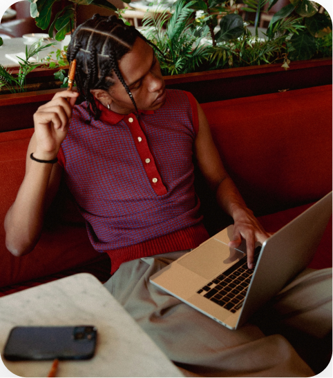 A stylish person with braids sits on a red booth in a cafe, looking thoughtful while holding a pencil to their head and typing on a laptop.