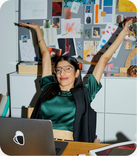 A joyful woman in a green silk top and glasses with her arms raised in celebration, sitting at a desk with a laptop and a mood board wall behind her.