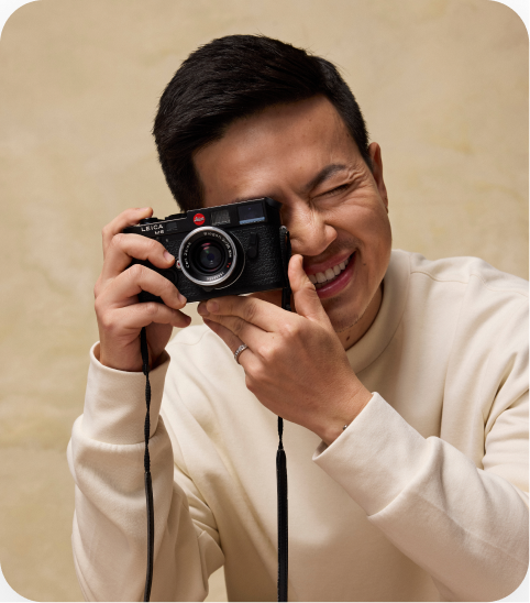 A smiling person in a cream-colored sweater taking a picture with a black film camera against a beige background.