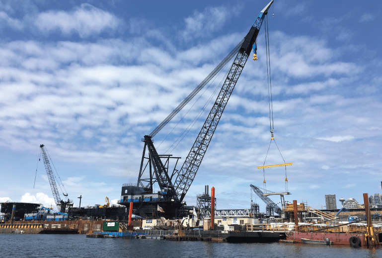 Large black crane lifting construction materials at a waterfront site with blue sky and scattered clouds.