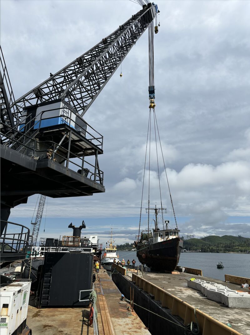 Crane lifting a fishing boat onto a dock with workers and water in the background under a cloudy sky.