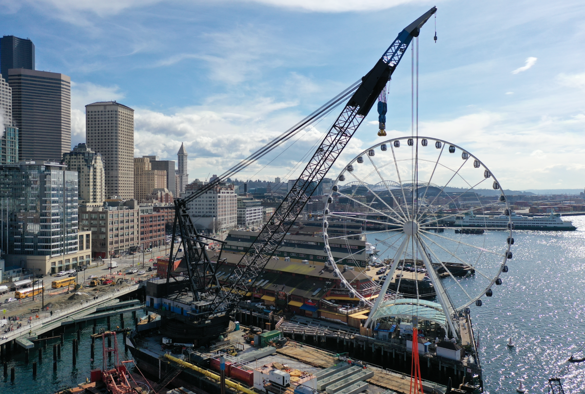 View of Seattle waterfront with a large crane, the Seattle Great Wheel, and city skyscrapers under a partly cloudy sky.