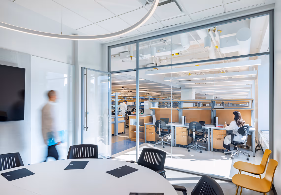 Modern office space showing a round conference table with chairs and a glass wall revealing workstations and a person in a white lab coat writing.