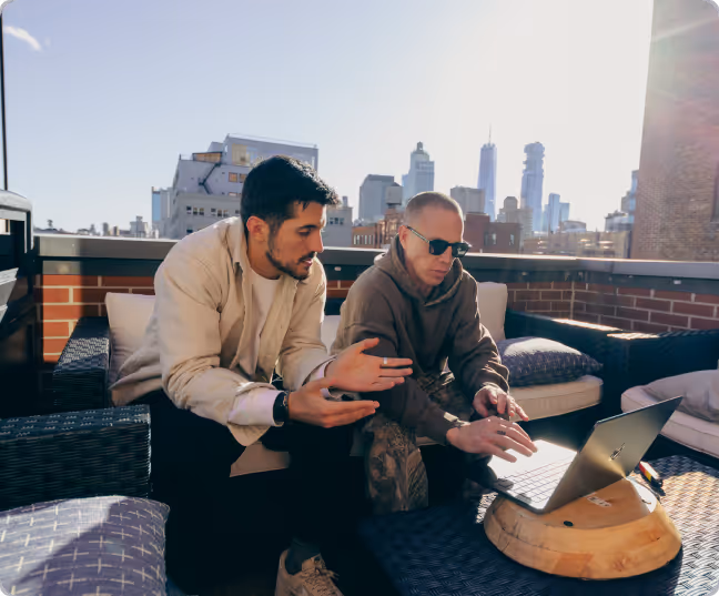 Ben and a man sitting on an outdoor patio sofa engaged in discussion while looking at a laptop on a low table with city skyscrapers in the background.