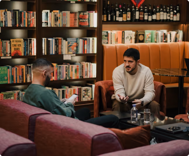 Ben and a man having a conversation in a cozy room with bookshelves and wine bottles in the background.