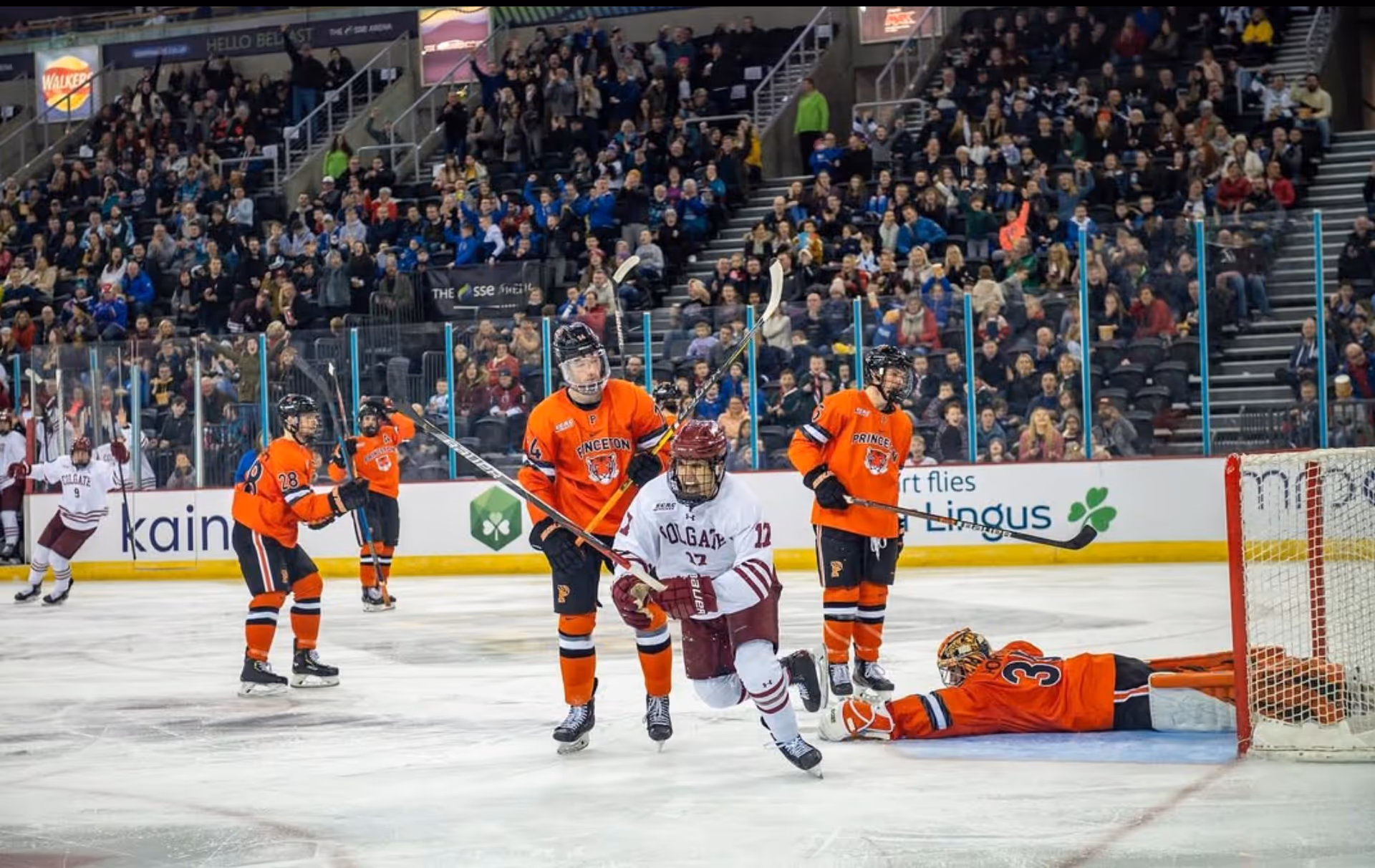 Hockey player from Colgate in white skates past Princeton players in orange near the goal, with crowd cheering in the background.