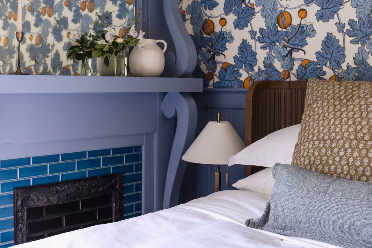 Bedroom corner with a blue fireplace, patterned wallpaper, wooden headboard, and pillows on a white bed.