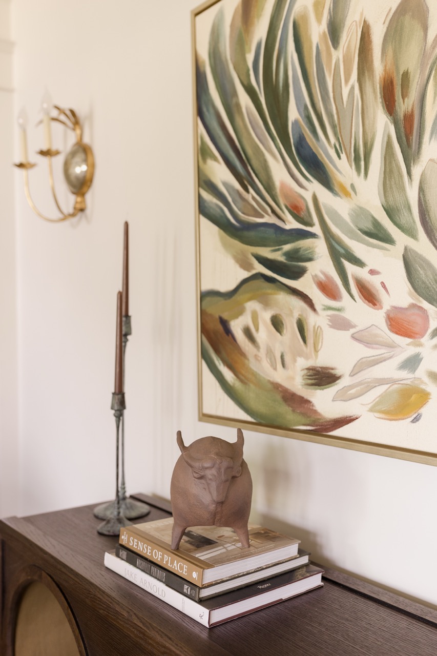 Decorative bull sculpture on stacked books on wooden cabinet under abstract framed painting.