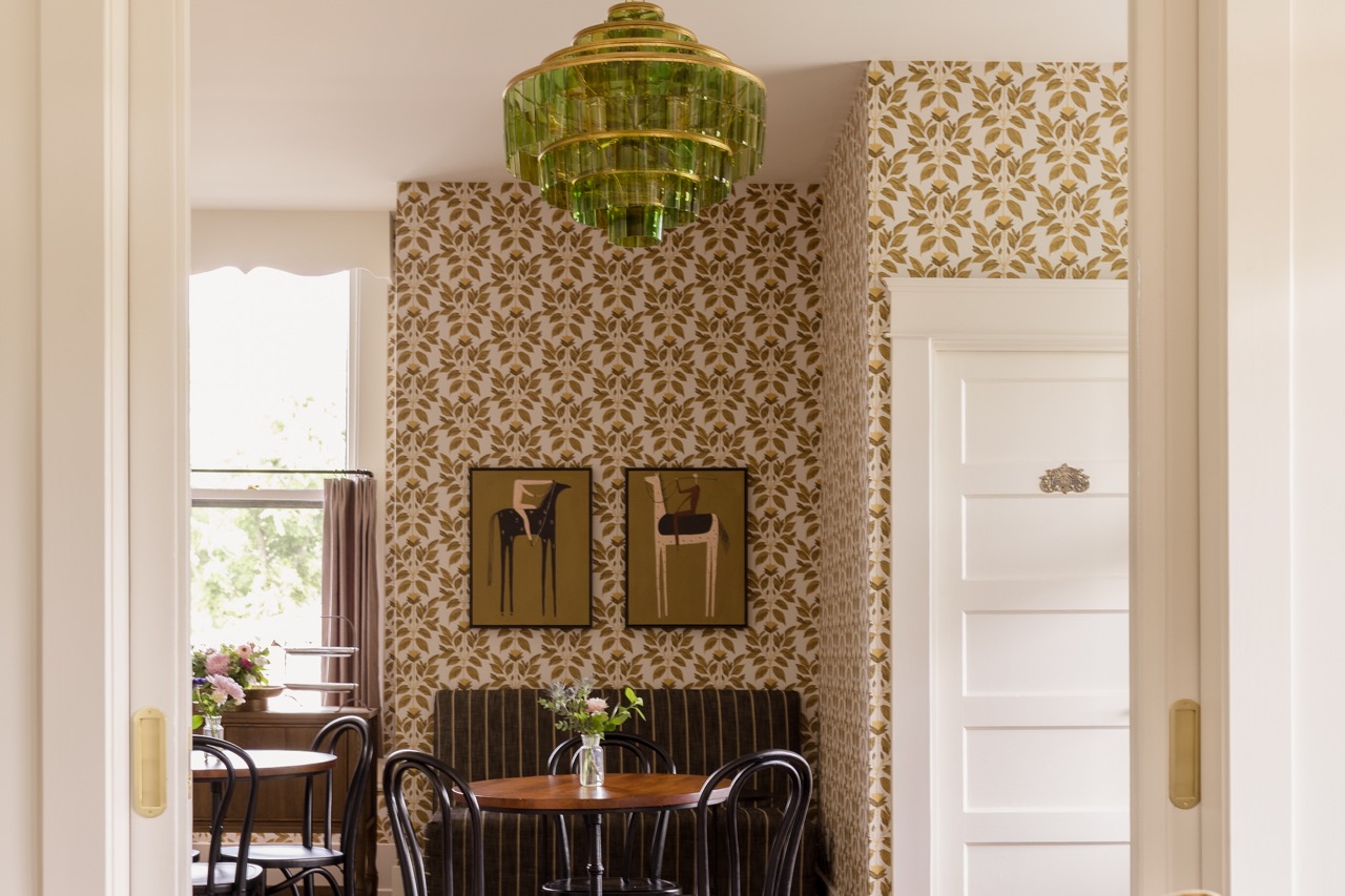 Cozy cafe corner with floral wallpaper, round wooden tables, black chairs, striped bench, and a green glass chandelier.