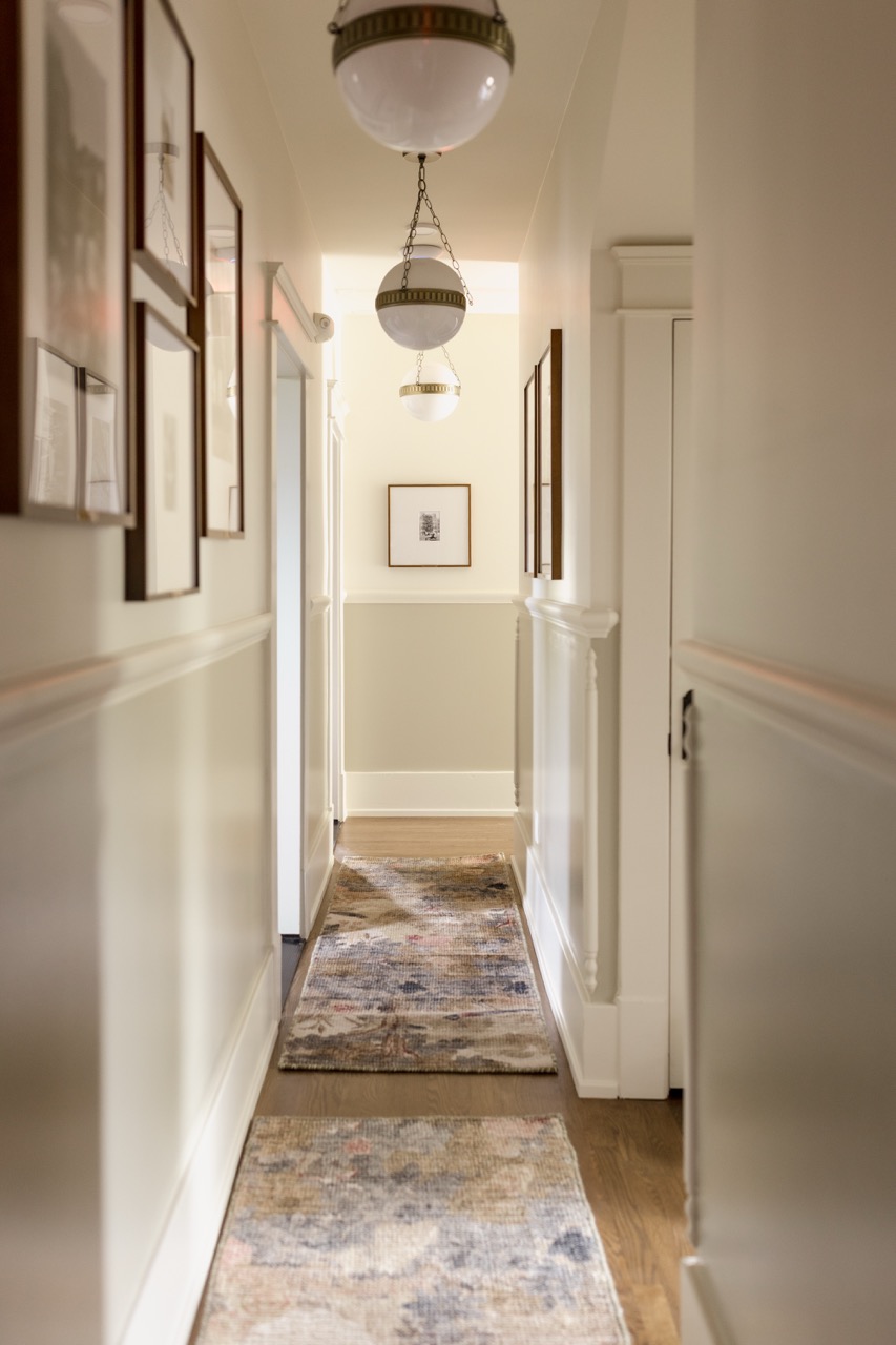 Narrow hallway with beige walls, three hanging globe lights, framed pictures, and patterned rugs on wooden floor.