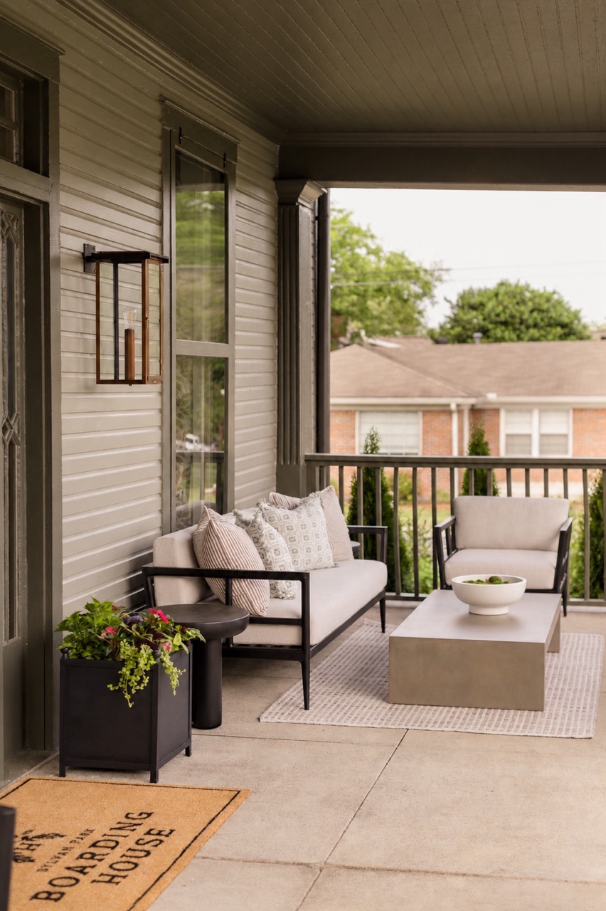 Cozy porch featuring beige cushioned sofa and chair with patterned pillows, coffee table, plant, and welcome mat.