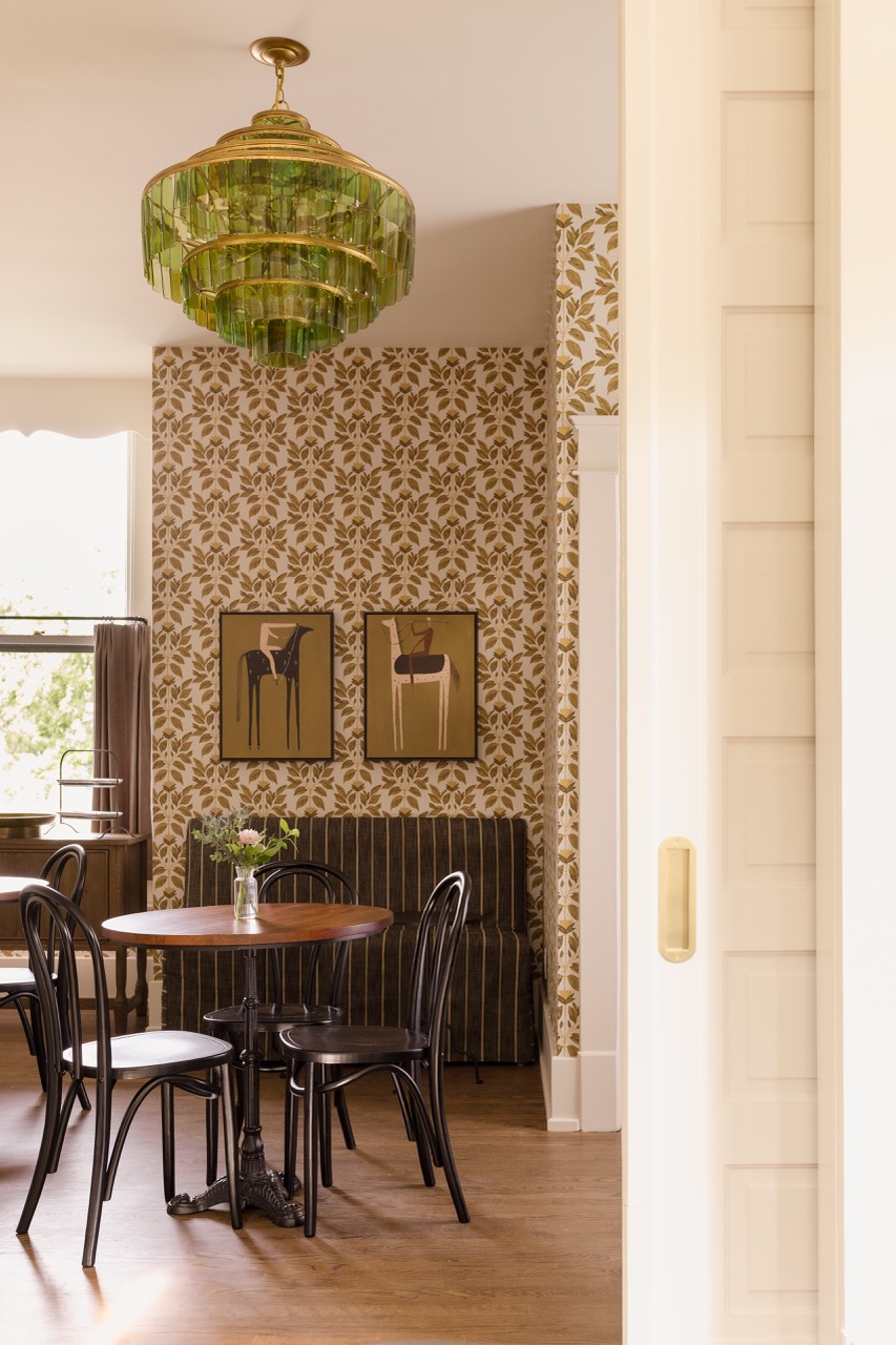 Cozy dining area with a round wooden table, four black chairs, floral wallpaper, and a green glass chandelier.
