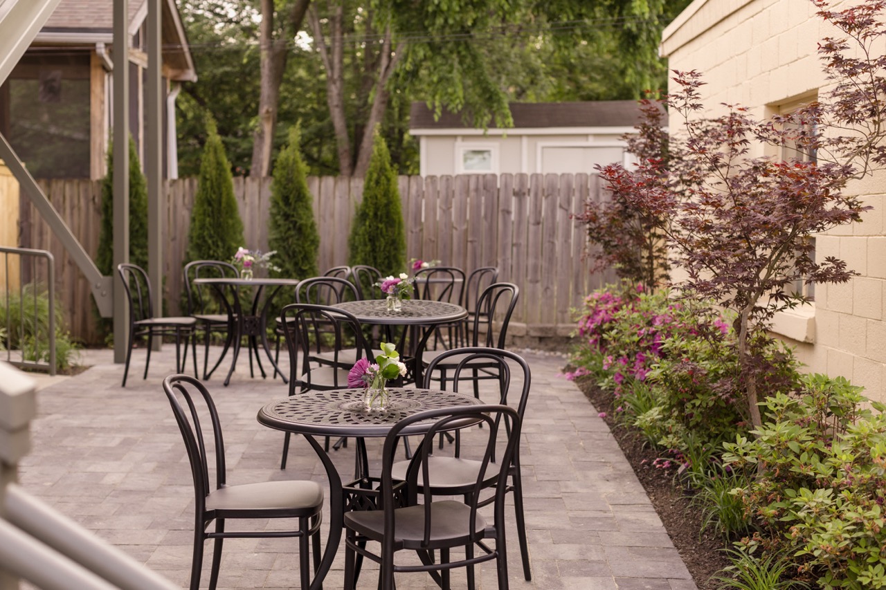 Outdoor courtyard with black metal tables and chairs, small flower vases on tables, and garden plants along a building.