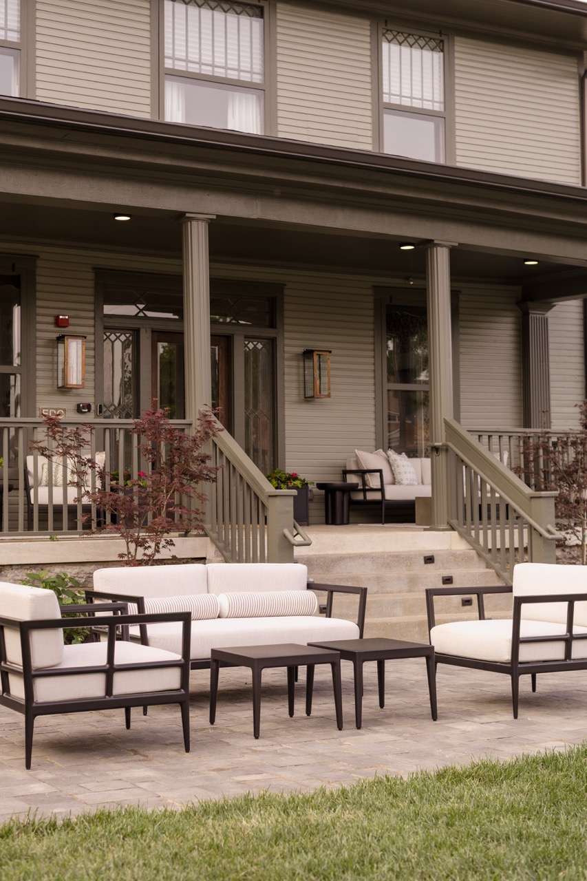 Outdoor seating area on a stone patio with white cushioned chairs and sofas in front of a house porch.