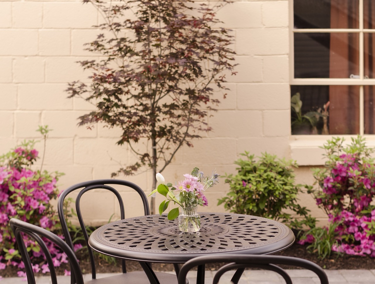 Metal patio table with a small vase of flowers in front of a beige brick wall and a window.