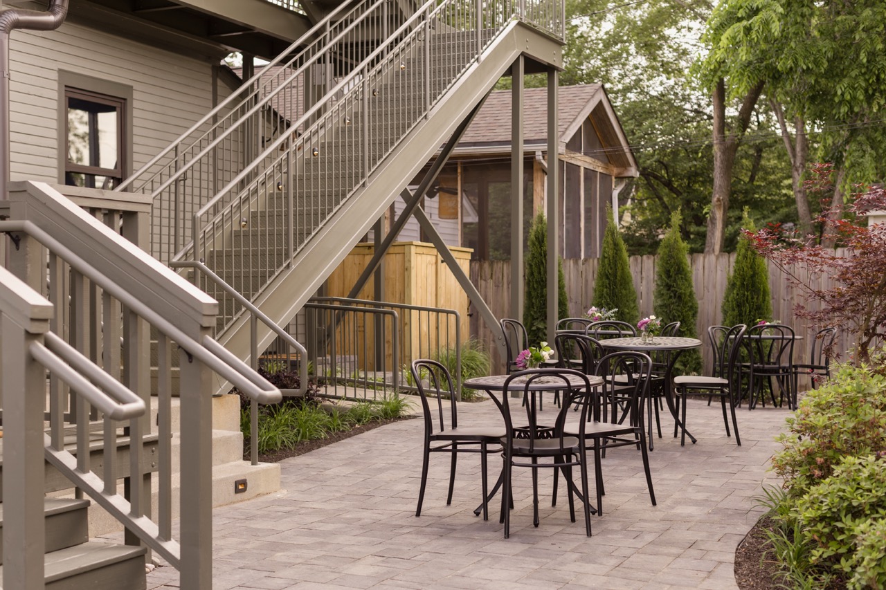 Outdoor courtyard with black metal tables and chairs, stone pavers, stairs, and greenery.