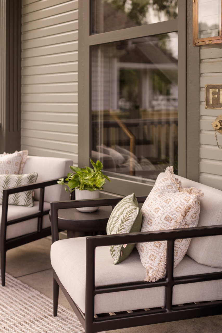 Front porch seating area with two cushioned chairs, decorative pillows, small round table, and potted plant.