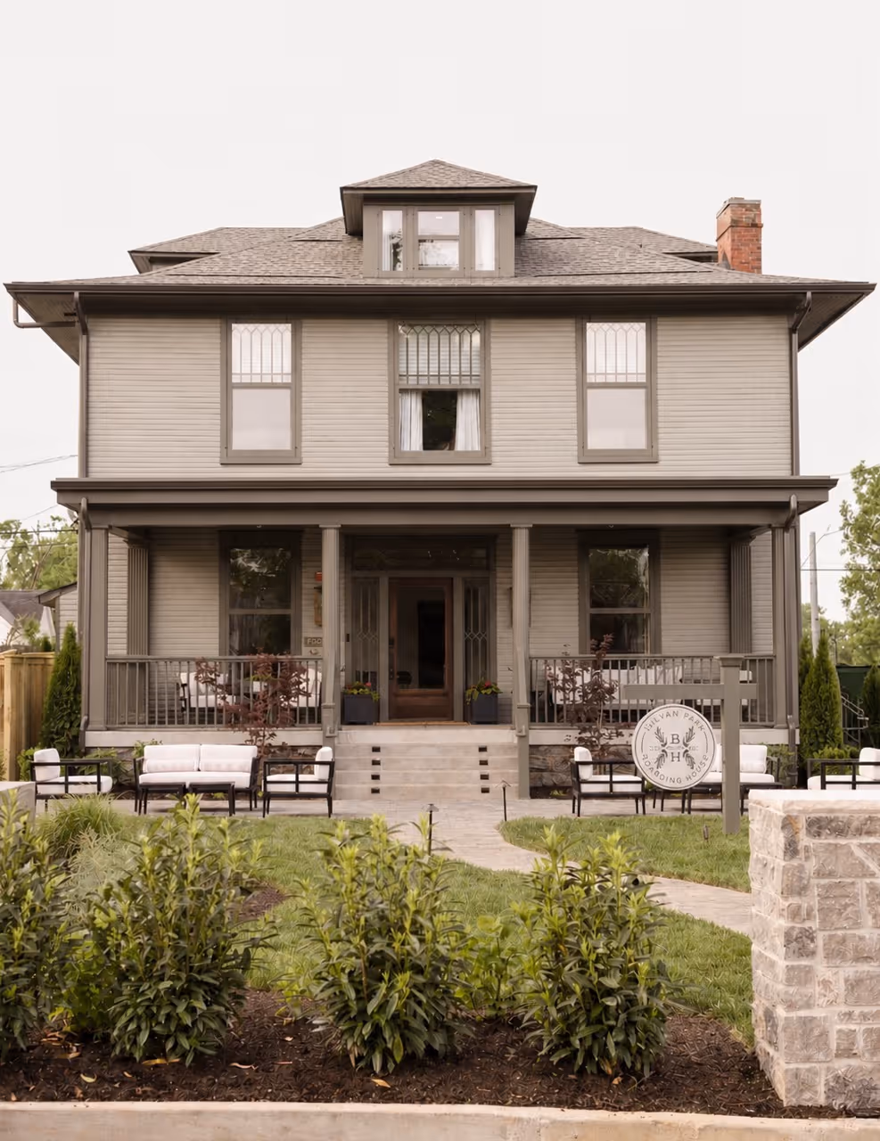 Two-story gray boarding house with front porch, outdoor seating, and landscaped garden path.
