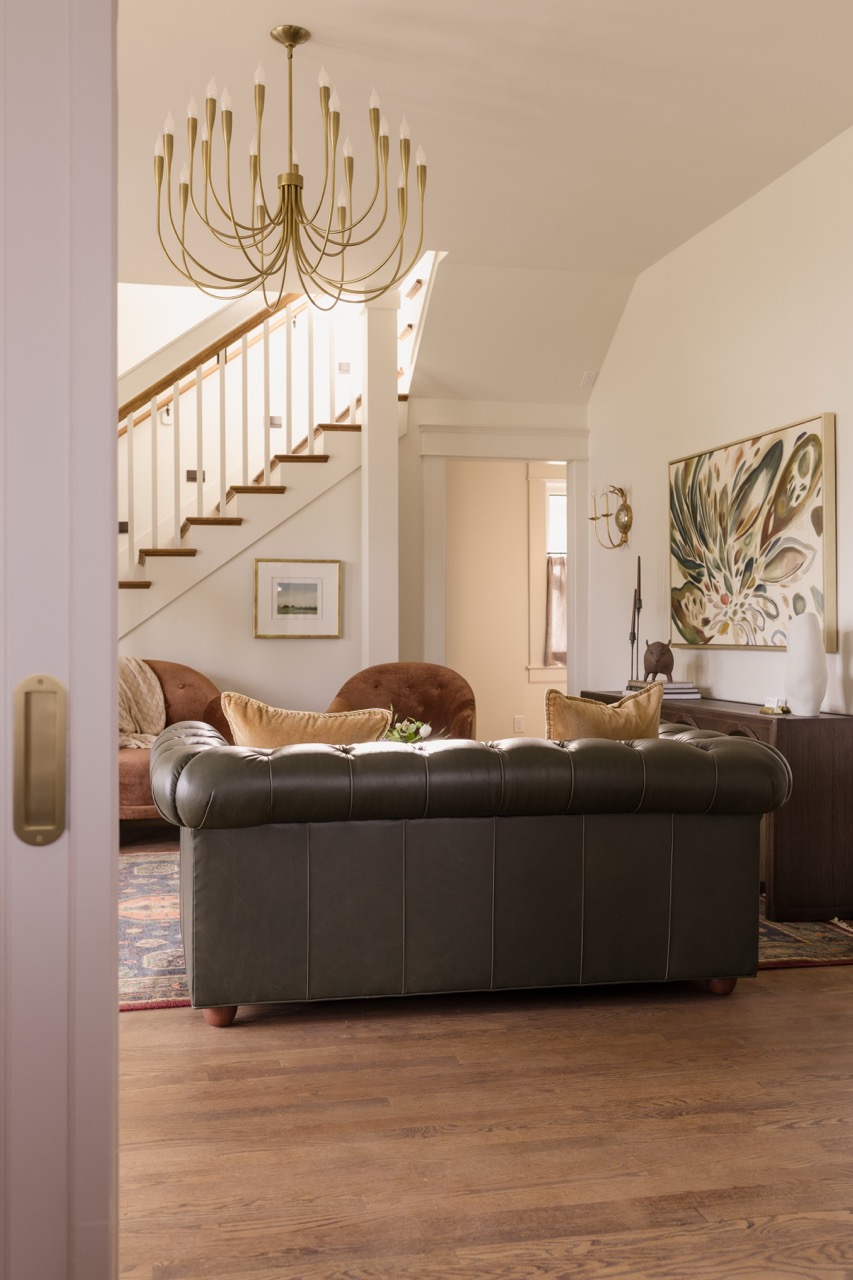 Living room with dark green leather sofa, brown chairs, abstract wall art, and a gold chandelier.