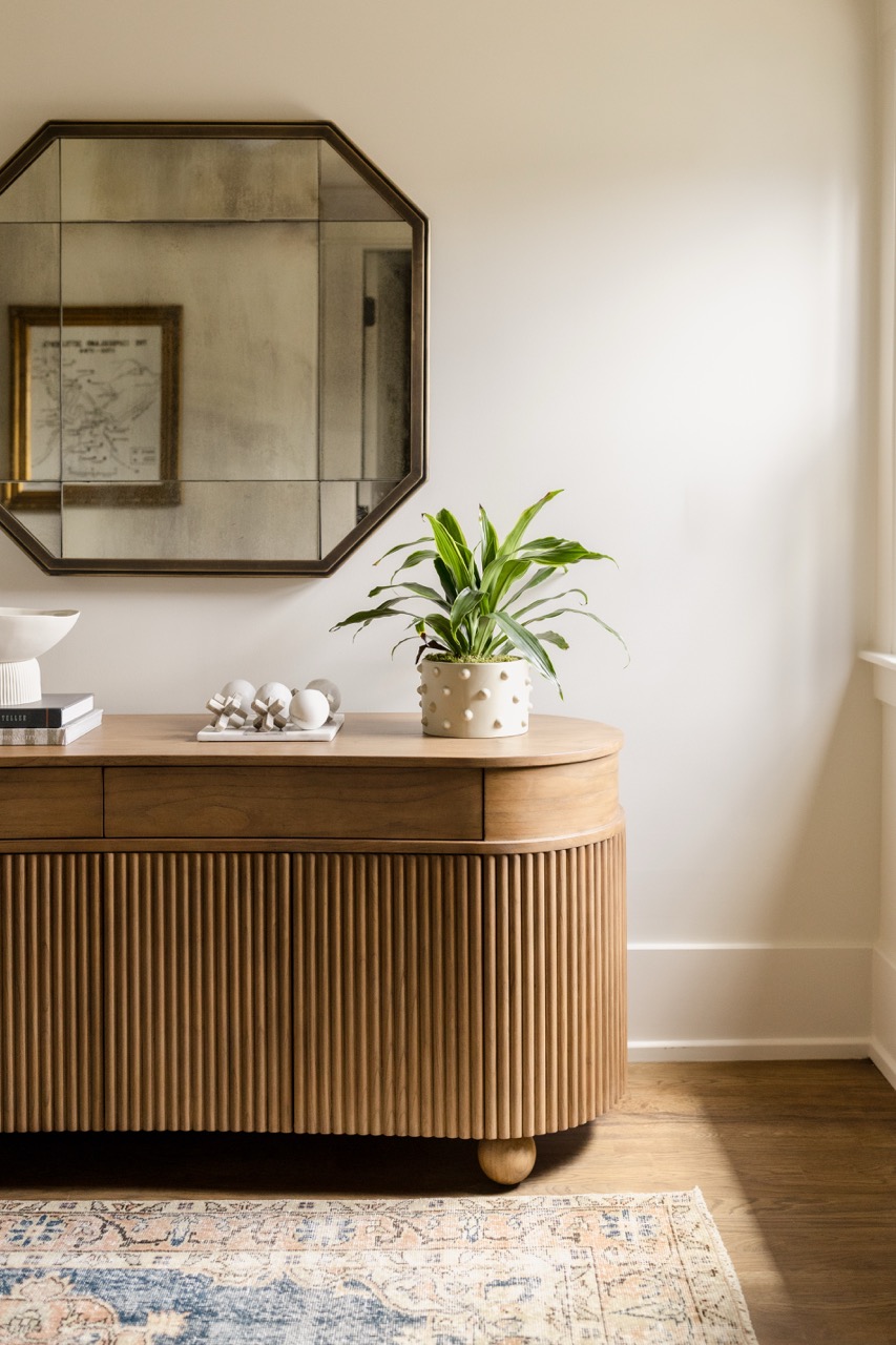 Wooden sideboard with ridged front panels holding a potted plant, decor items, and books under an octagonal mirror.