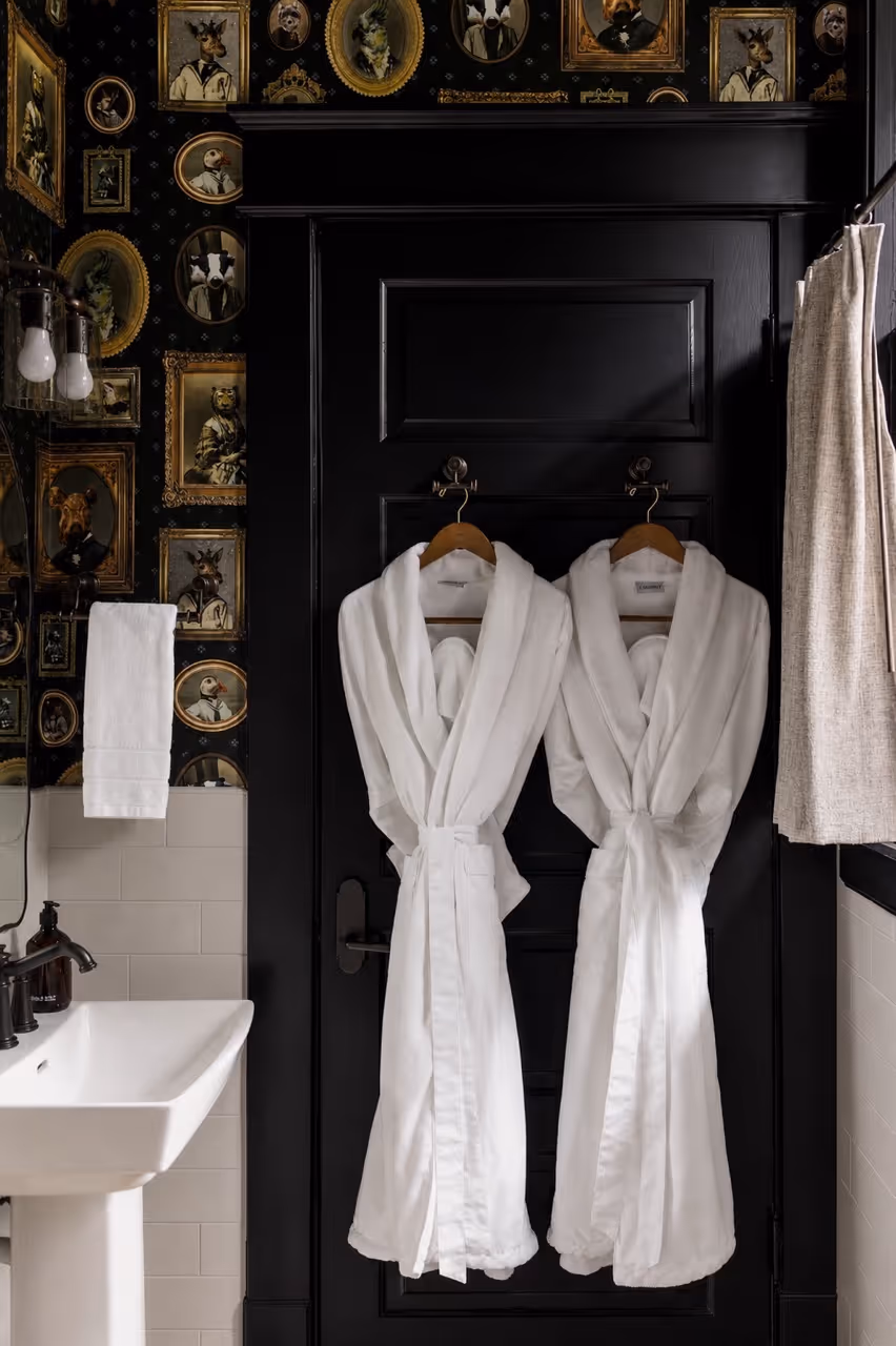 Two white bathrobes hanging on a black door in a bathroom with animal portrait wallpaper.