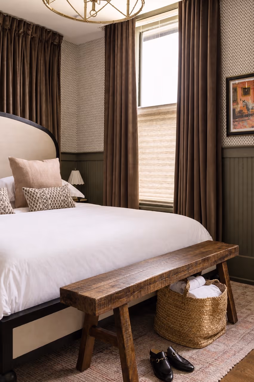 Cozy bedroom corner with a bed, wooden bench, woven basket with towels, and window with brown curtains.
