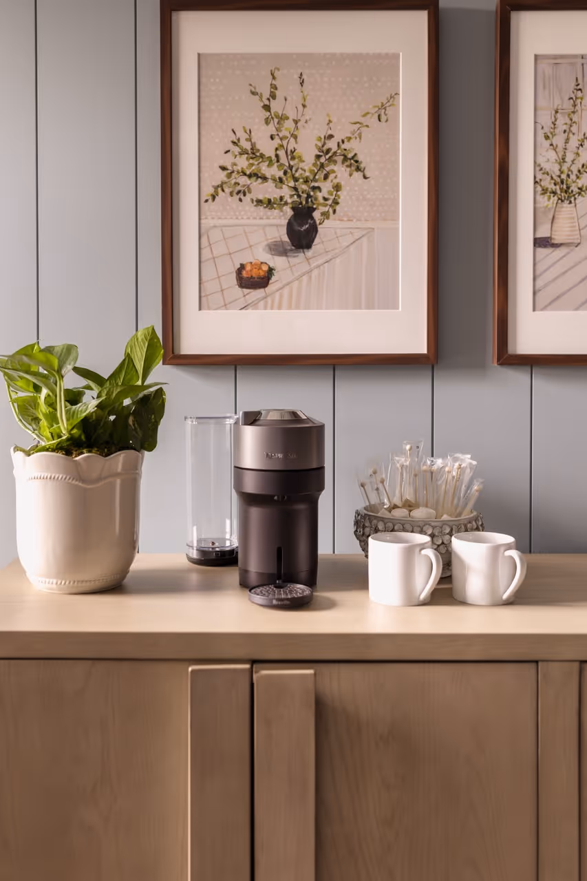Black coffee grinder on a wooden cabinet with two white mugs, a potted plant, and framed botanical prints on a grey wall.