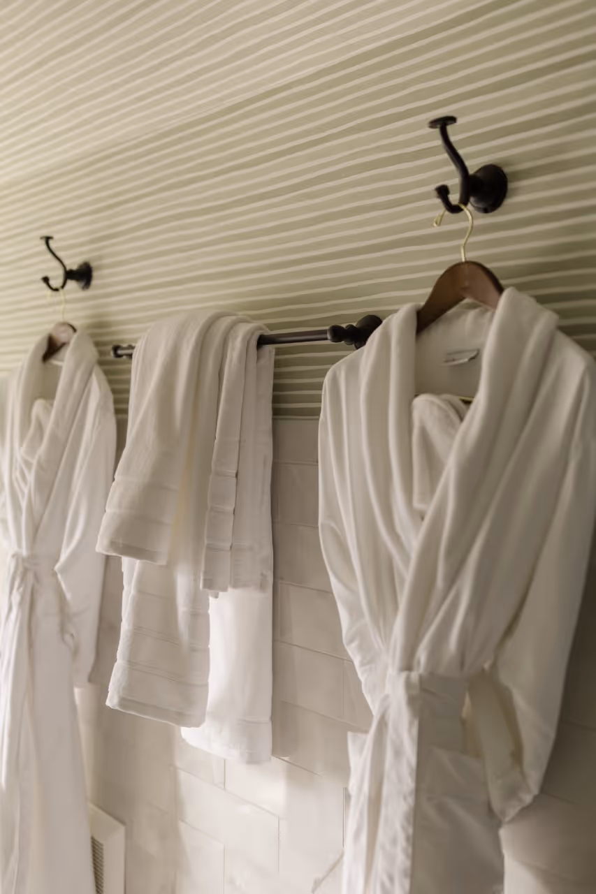 White bathrobes hanging on hooks and white towels draped on a towel rack in a striped wallpapered bathroom.