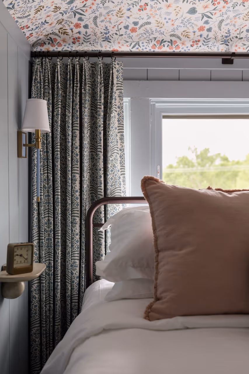 Corner of a bed with white linens, a large pink pillow, floral curtains, and a floral wallpapered ceiling.
