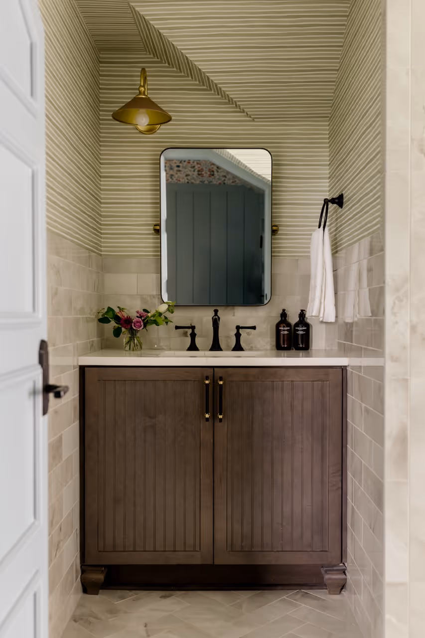 Small bathroom vanity with dark wooden cabinet, white countertop, black faucet, rectangular mirror, and striped wallpaper.