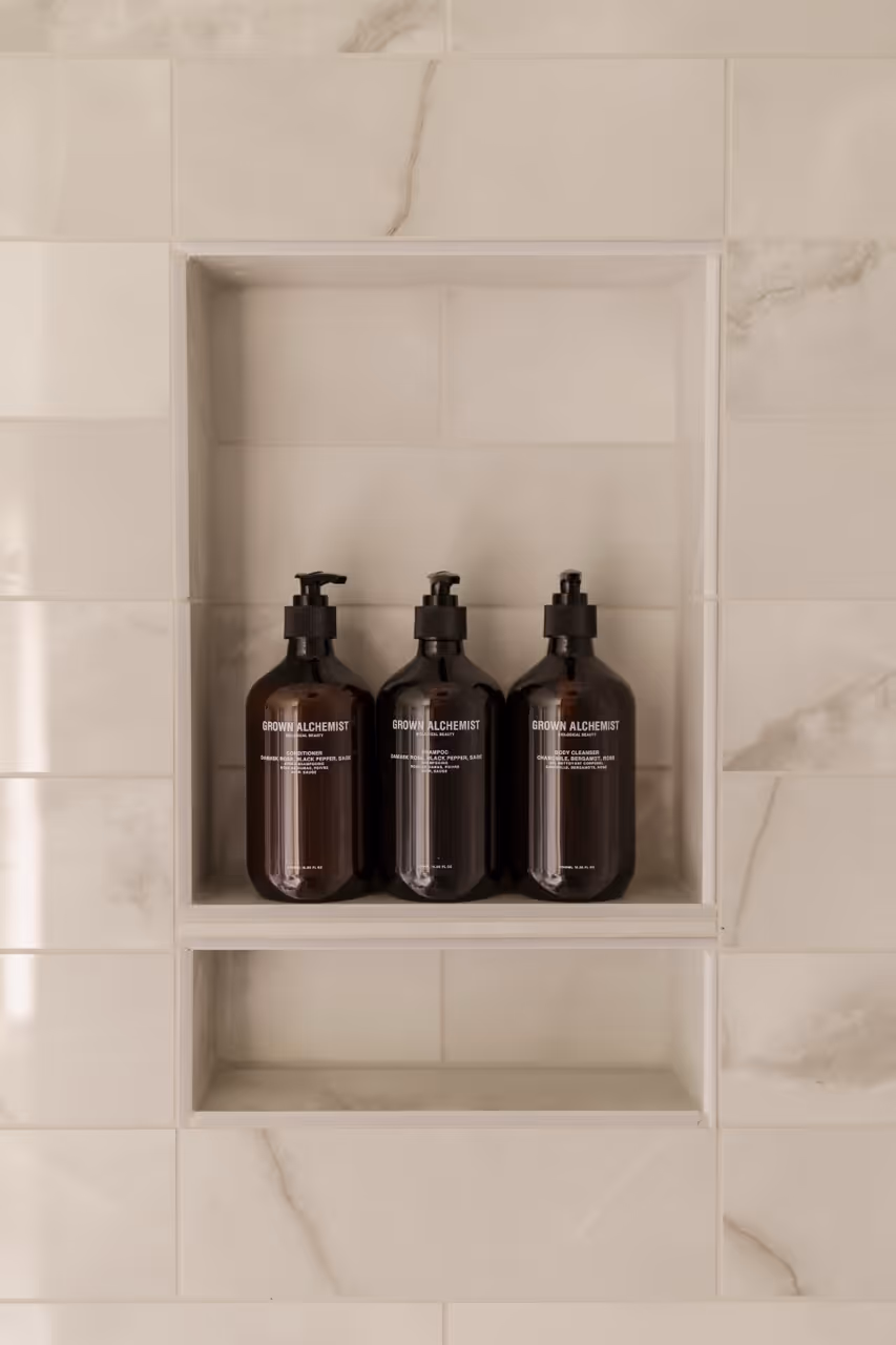 Three Grown Alchemist toiletry bottles with pumps inside a recessed shower shelf with cream marble tiles.