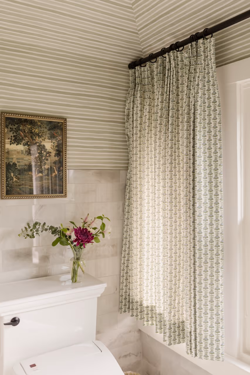 Bathroom corner with white toilet, window with green leaf-patterned curtain, and small vase of flowers.