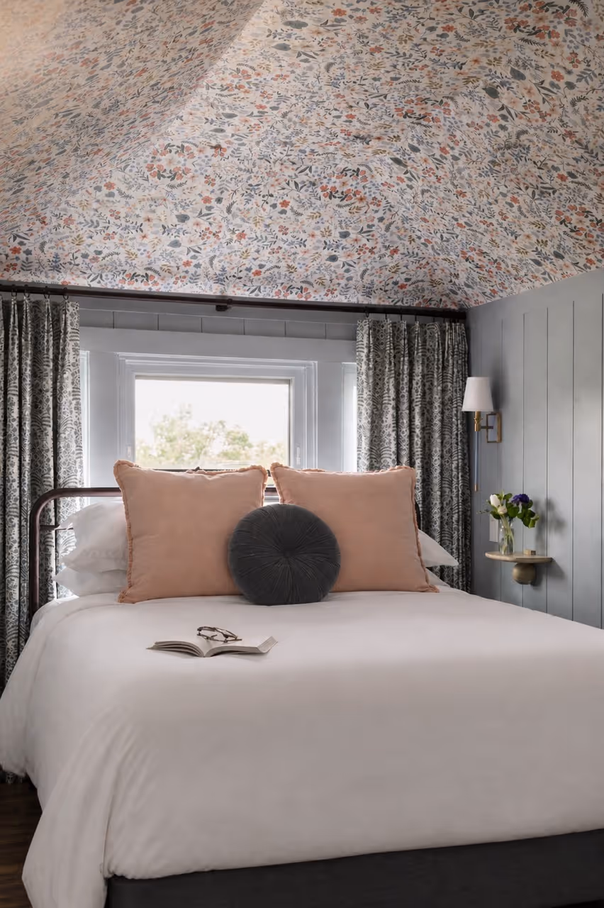 Cozy bedroom with a floral wallpapered ceiling, two beige pillows, and a round gray cushion on a white bed.