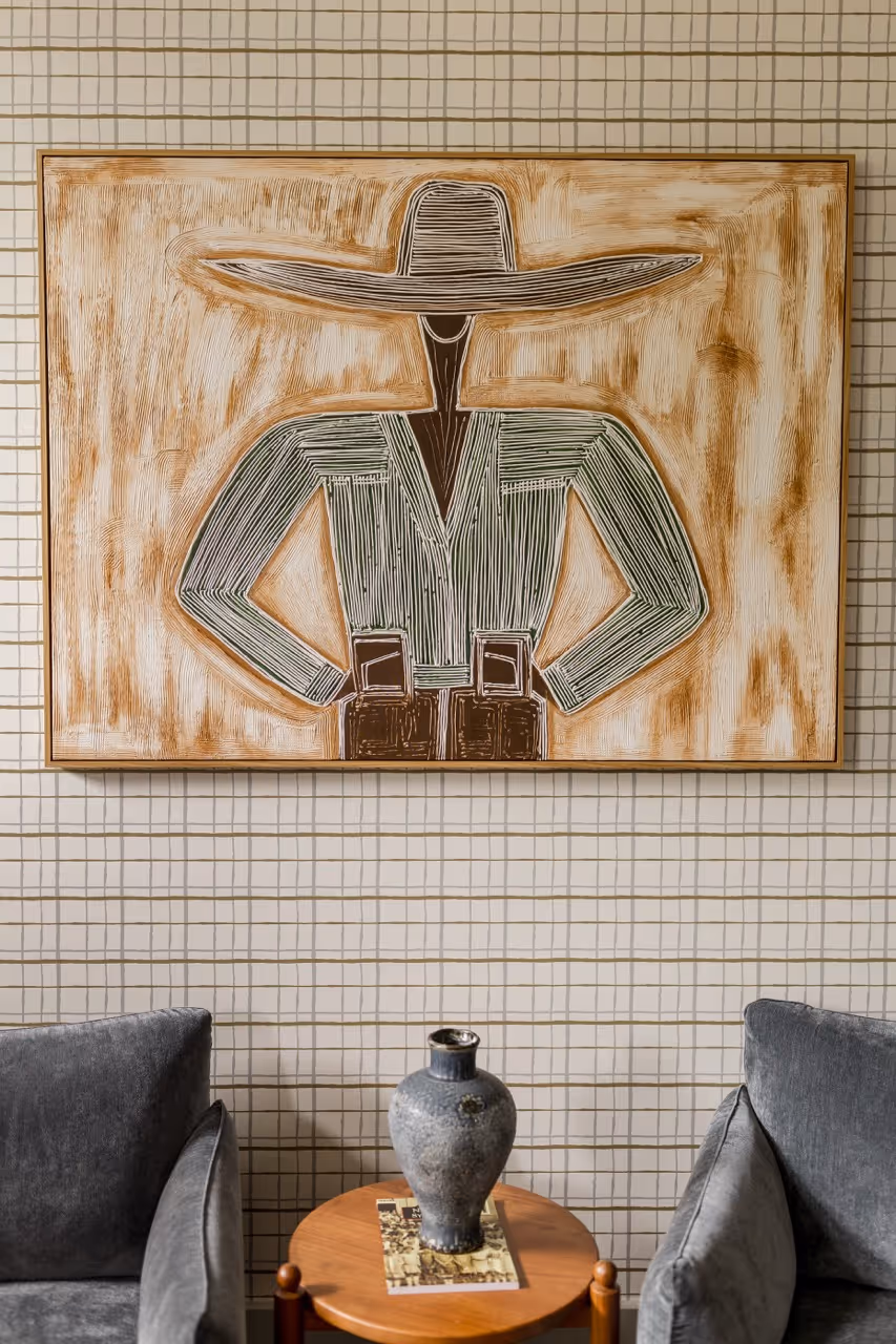 Abstract artwork of a person wearing a wide-brimmed hat hanging above two grey armchairs and a wooden table with a vase.