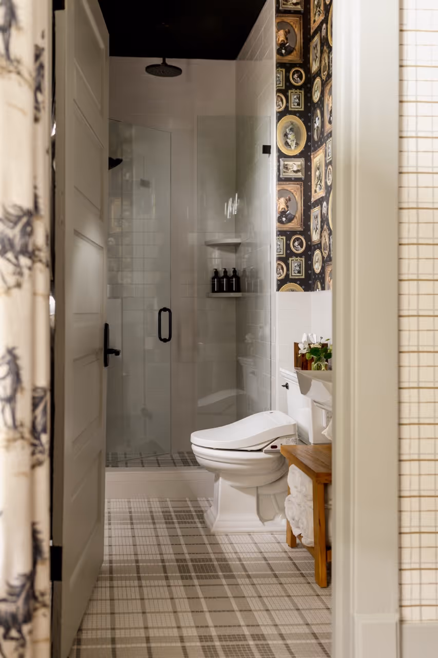 Bathroom with glass shower, white toilet, patterned floor, and wallpaper featuring portrait frames.