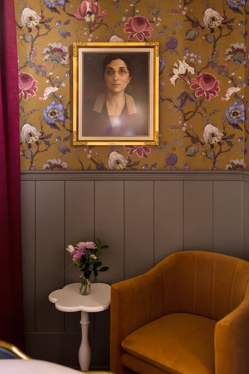 Vintage-style room corner with floral wallpaper, framed portrait, mustard armchair, and small white table with flowers.