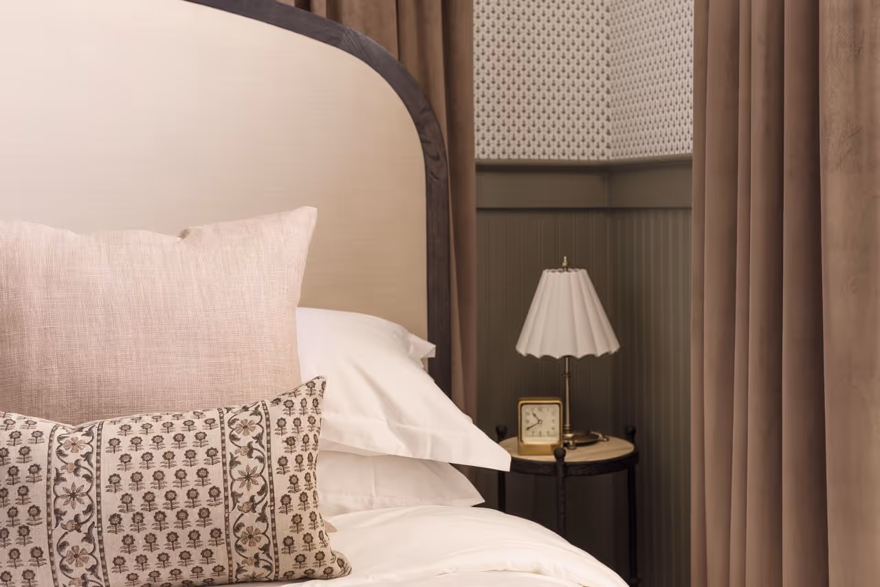 Cozy bedroom corner with beige padded headboard, decorative pillows, bedside table with lamp and clock.